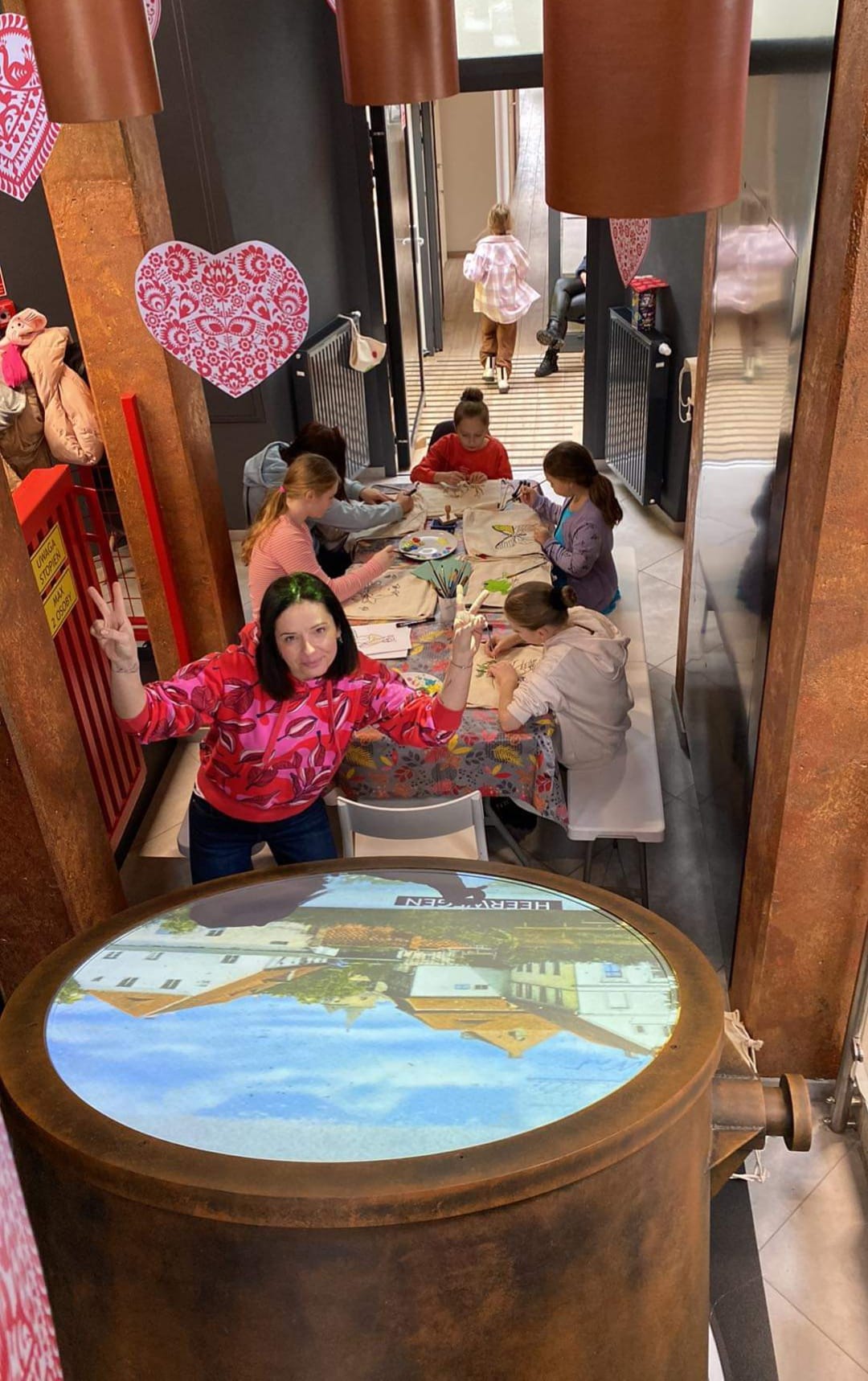 Kobieta przed ekranem w kształcie koła, w tle dzieci przy stoliku, malujące na torbach
