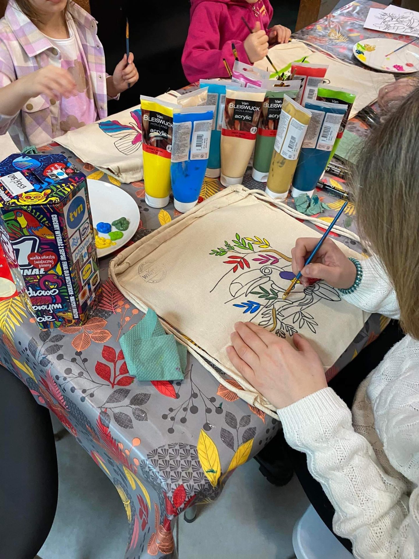 Dziewczynka malująca na torbie tukana, w tle tubki z farbami i puszka WOŚP